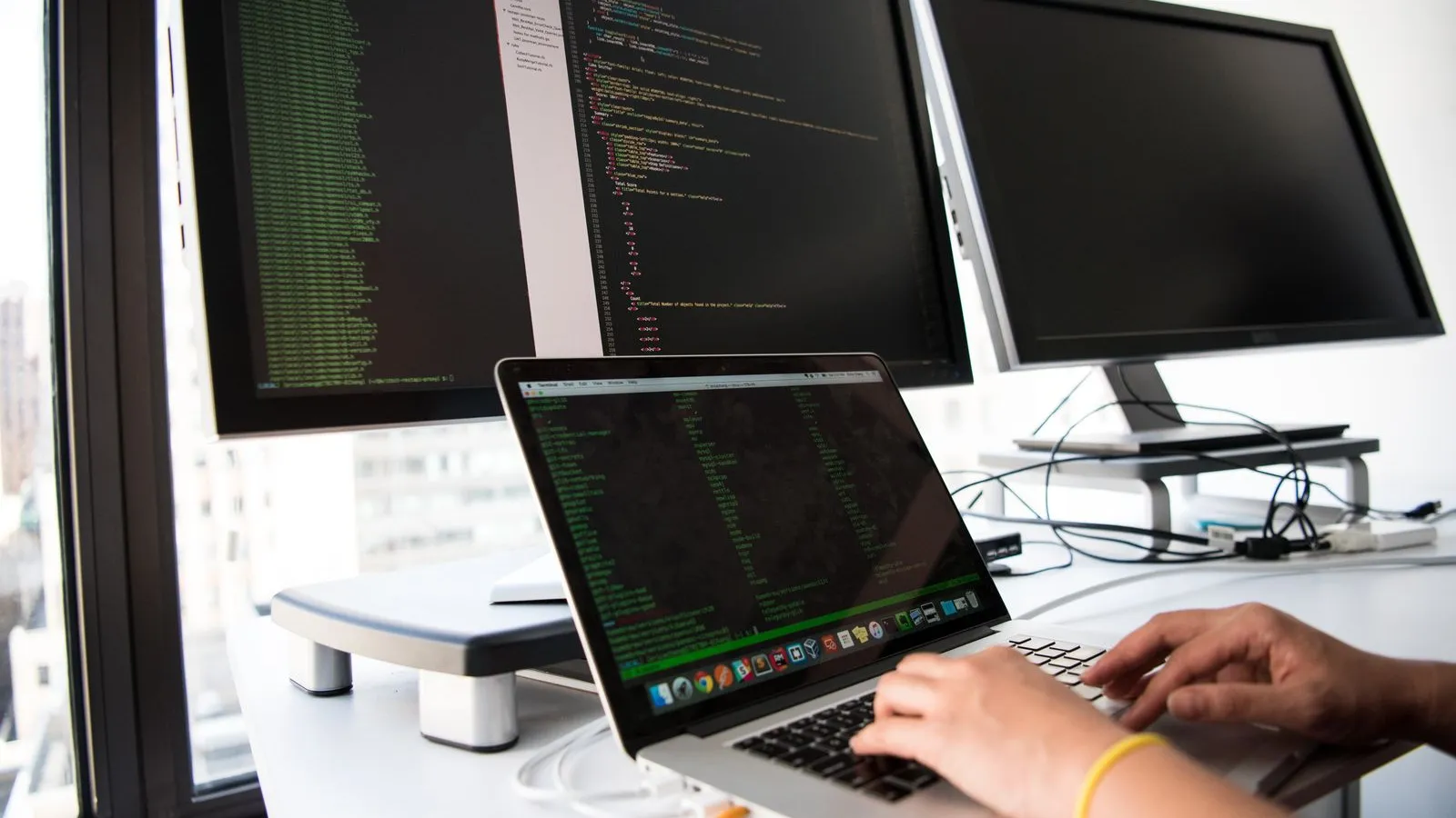 Programátor pri notebooku a dvoch monitoroch s kódom – tvorba webovej aplikácie na mieru